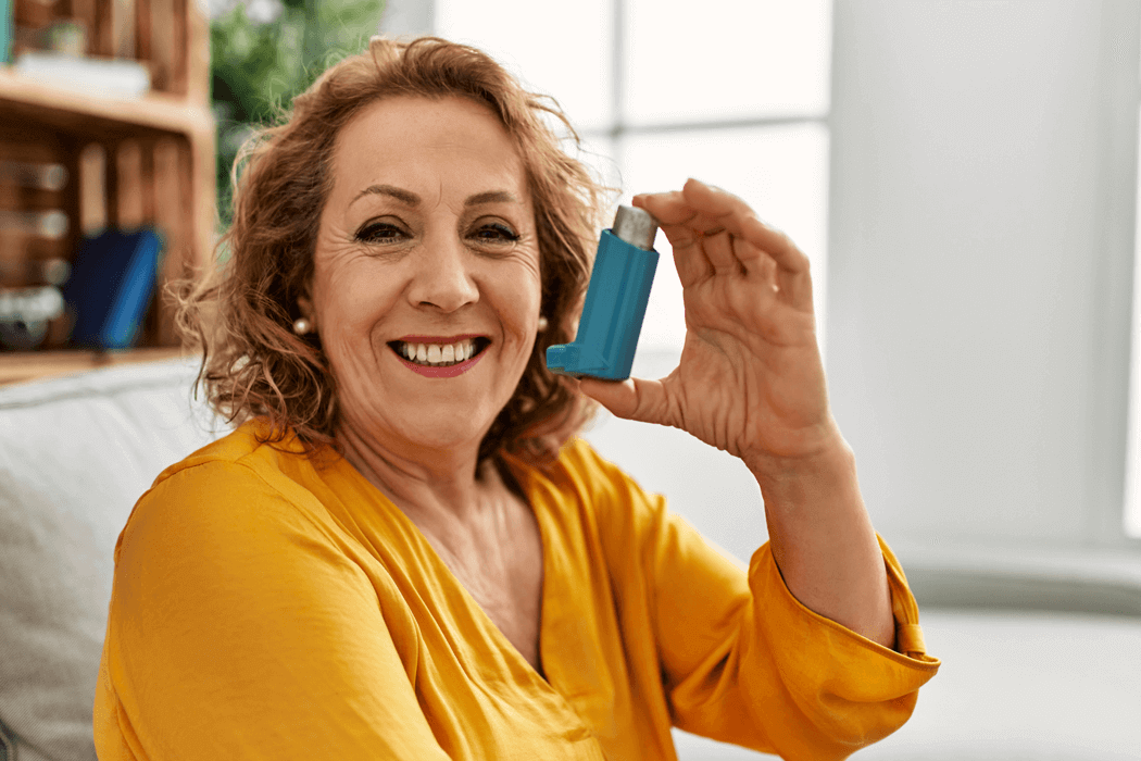 Older woman with curly hair and a yellow top holding up an inhaler while smiling