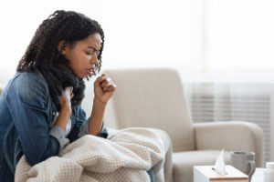 A sick Black woman is coughing intensely at home, displaying symptoms of a respiratory virus.