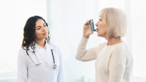 A physician is examining a mature woman who is using a blue asthma inhaler.