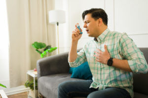 Latin man having breathing problems using an inhaler pump for asthma and touching his chest because of the pain