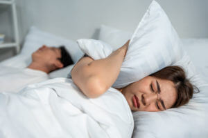 A woman lies awake in bed looking frustrated as her husband snores loudly beside her due to sleep apnea, disrupting her sleep.